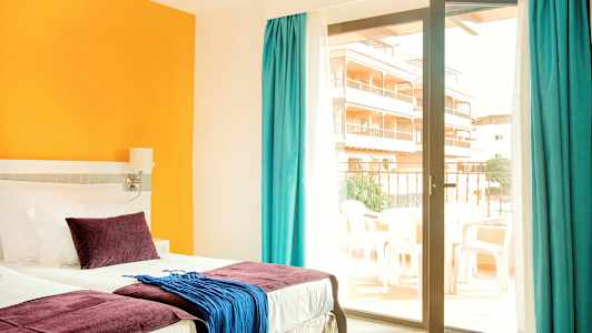 Ett hotellrum med en säng, färgglada kuddar och textilier, samt en glasdörr som leder till en balkong. (AI-genererad text)