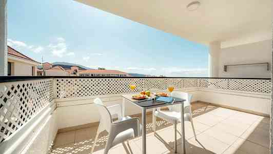 Balkong med bord och stolar samt utsikt över omgivande byggnader och berg. (AI-genererad text)