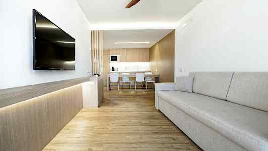 Ett modernt vardagsrum med ljus inredning, en grå soffa, en väggmonterad TV och ett kök i bakgrunden. (AI-genererad text)