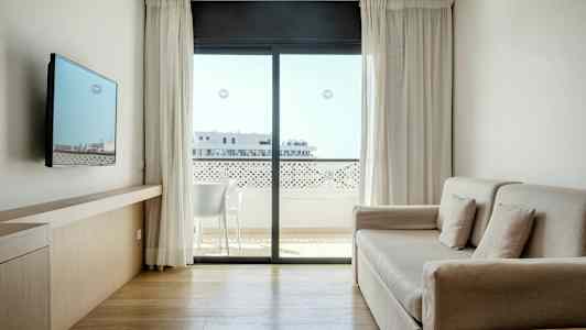 En ljus och modern hotellrum med soffa, väggmonterad TV och stort fönster med utsikt mot en balkong. (AI-genererad text)