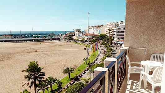 Vy från en balkong med bord och stolar mot en sandstrand, grönskande park och stadsmiljö. (AI-genererad text)