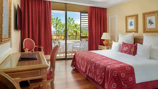 Ett hotellrum med en stor säng, sittmöbler och en balkong med utsikt över palmer. (AI-genererad text)