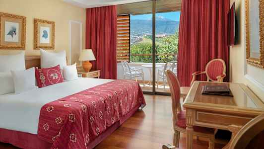 Hotellrum med dubbelsäng, skrivbord, stol och balkong med utsikt mot grönska och berg. (AI-genererad text)