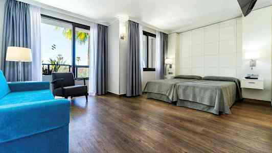 Ett hotellrum med två enkelsängar, en blå soffa, en fåtölj och stora fönster med utsikt. (AI-genererad text)