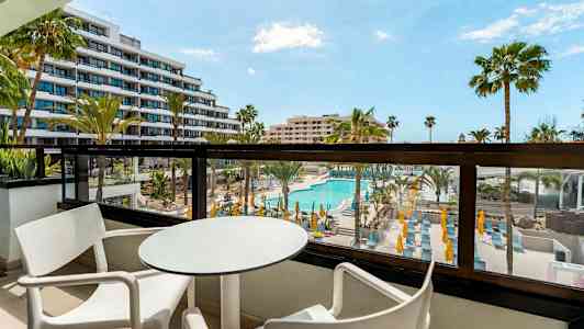 Balkong med bord och stolar, utsikt över poolområde och omgivande byggnader. (AI-genererad text)