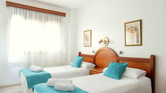 Två enkelsängar med turkosa prydnadskuddar och vitt överkast i ett soligt rum med en stor fönsterruta. (AI-genererad text)