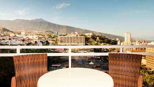 Ett bord och två stolar på en balkong med utsikt över en stad och ett berg i bakgrunden. (AI-genererad text)