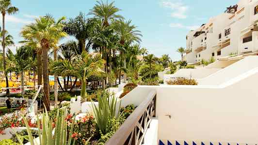 Utsikt över en trädgård med flera vajande palmträd och vita byggnader med terrasser. (AI-genererad text)