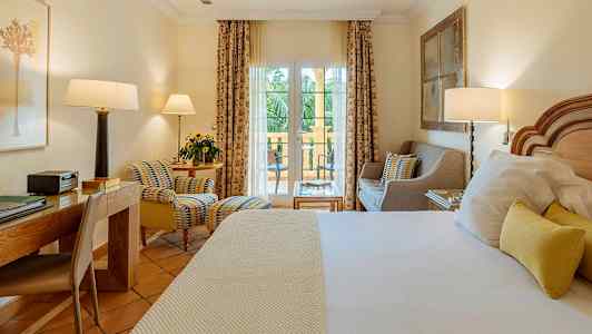 Hotellrum med säng, skrivbord och sittgrupp framför fönster som leder till balkong. (AI-genererad text)