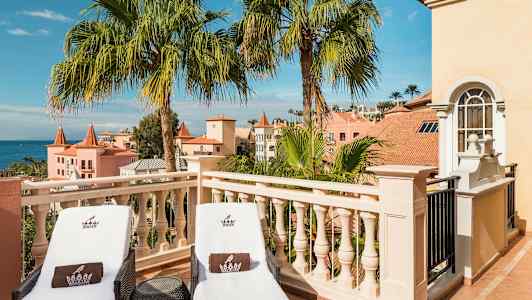 Två solstolar på en terrass med utsikt över palmer och pastellfärgade byggnader. (AI-genererad text)