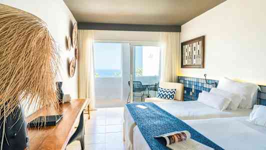 Ett ljust hotellrum med två bäddar, en träbänk och en balkong med havsutsikt. (AI-genererad text)