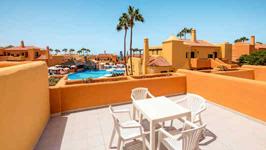 En terrass med bord och stolar, med utsikt över en pool och omgivande byggnader. (AI-genererad text)