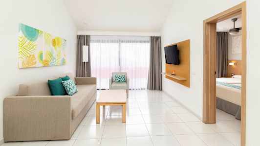 Ett hotellrum med en delvis synlig bädd, en soffa och fåtölj samt ett bord. Ett fönster med gardiner syns i bakgrunden. (AI-genererad text)