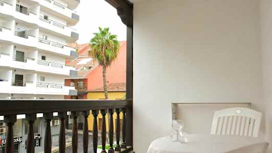 En balkong med ett bord, två glas och en vit plaststol. Utsikten visar byggnader, en palm och en gata. (AI-genererad text)