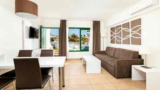 Ett ljus och modernt inrett hotellrum med en brun soffa, ett matbord och stolar. Fönster och dörr leder till utomhusområdet. (AI-genererad text)