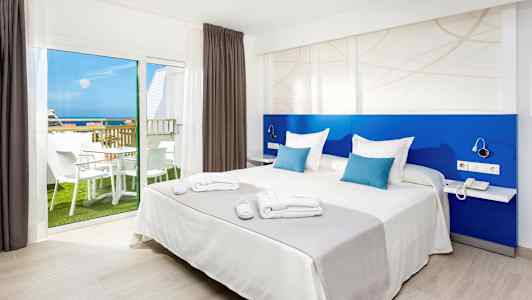 Bild av ett hotellrum med en dubbelsäng, blå ryggstöd och ett fönster som visar en balkong med möblemang och havsutsikt. (AI-genererad text)