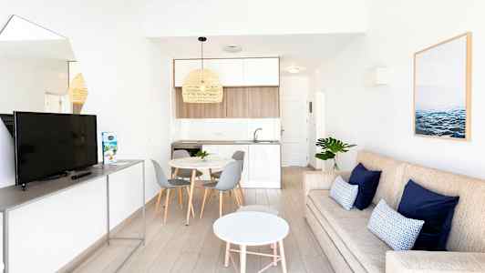 Ett modernt vardagsrum med en beige soffa, blå kuddar, en vit rund soffbord och en tv på ett stativ. Bredvid finns ett matområde med ett runt bord och grå stolar samt ett kök i ljusa färger. (AI-genererad text)