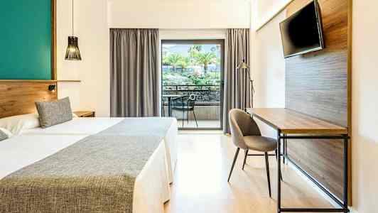 Hotellrum med två sängar, skrivbord, TV och balkong med utsikt över träd och grönska. (AI-genererad text)