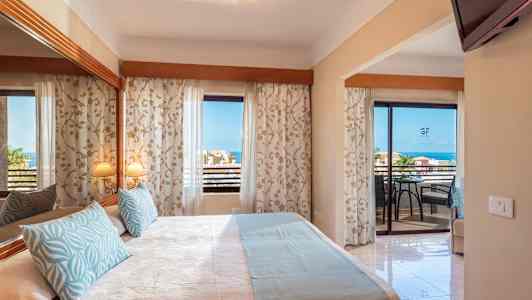 Ett hotellrum med en dubbelsäng, dekorativa kuddar och en spegel. Balkong med utsikt mot havet. (AI-genererad text)