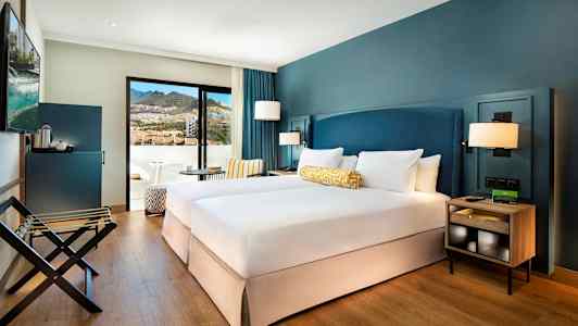 Ett hotellrum med två enkelsängar, blå väggar och en balkong med utsikt över bergen. (AI-genererad text)