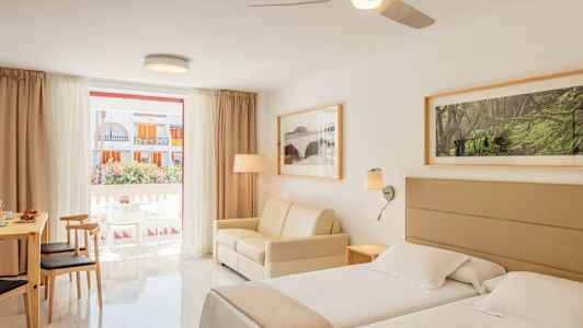 Ett ljust hotellrum med två sängar, en soffa och matsalsbord med stolar, samt ett stort fönster med utsikt över balkongen och byggnader utanför. (AI-genererad text)