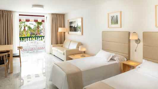 Hotellrum med två enkelsängar, en soffa, ett bord med stolar och ett fönster mot en balkong. (AI-genererad text)