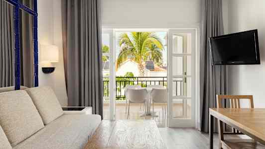 Ett vardagsrum med stor soffa, träbord och en TV, öppnar upp mot en balkong med utsikt över tropiska träd och byggnader. (AI-genererad text)