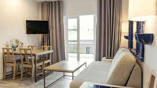 Ett vardagsrum med en grå soffa, ett träbord med stolar, en väggmonterad TV och glasdörrar med gardiner. (AI-genererad text)