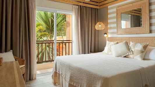 Ett hotellrum med en dubbelsäng, ljusa textilier och inredningsdetaljer. Utanför syns en balkong med utsikt över grönska. (AI-genererad text)