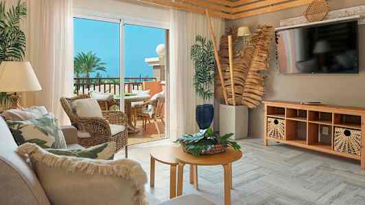 Ett vardagsrum med trädetaljer och soffmöbler, utgång till en möblerad balkong med utsikt över palmer och spirande vegetation. (AI-genererad text)