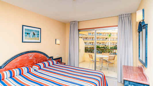 Hotellrum med två enkelsängar, randiga blå, röda och vita överkast, och utsikt mot en balkong med möbler. (AI-genererad text)