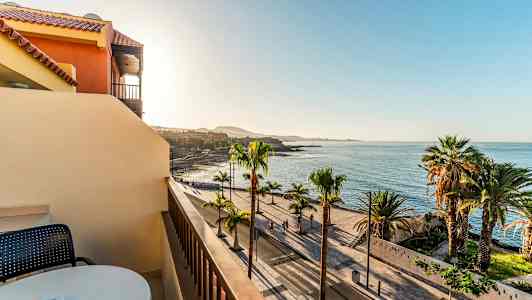 Utsikt från en balkong mot en palmkantad strandpromenad och havet under en klar himmel. (AI-genererad text)