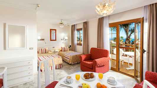 Ett hotellrum med två sängar, en röd fåtölj och en matplats, samt en balkong med utsikt över palmer och havet. (AI-genererad text)