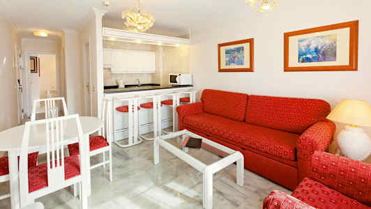 Ett vardagsrum med en soffa och fåtöljer klädda i röd textil, ett glasbord och ett kombinerat kök samt matplats med vita möbler. (AI-genererad text)