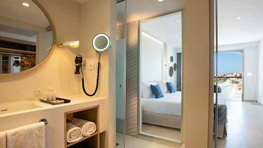 Hotellrum med en spegel och handfat bredvid ett sovrum med utsikt. (AI-genererad text)