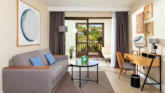 Ett hotellrum med en grå soffa, ett skrivbord och en balkong med utsikt över en trädgård. (AI-genererad text)