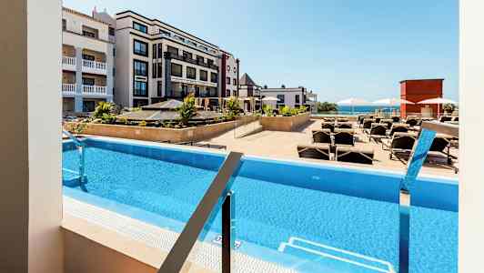 Utsikt över en pool med solstolar, omgiven av hotellbyggnader. (AI-genererad text)