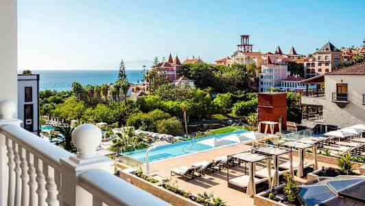 Utsikt från balkong över hotellområdet med pool och trädgård samt havet i bakgrunden. (AI-genererad text)