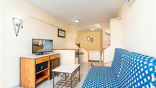 En studio med en soffgrupp, ett bord, samt en byrå med en TV på. I bakgrunden finns en säng. (AI-genererad text)