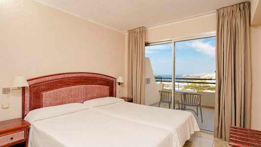 Ett hotellrum med två enkelsängar, ett trähuvudgavel och nattduksbord. Ett glasparti med utsikt mot havet och balkongmöbler. (AI-genererad text)