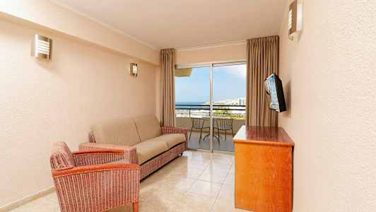 Ett vardagsrum med en soffa, en fåtölj, ett träbord och en väggmonterad TV. I bakgrunden finns en balkong med stolar och utsikt mot havet. (AI-genererad text)