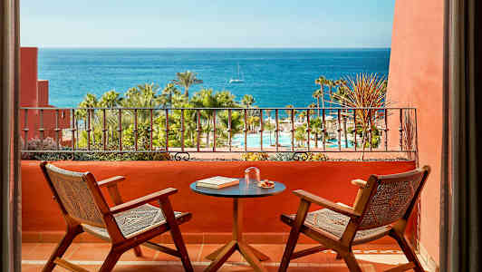 Två stolar och ett bord på en terrass med utsikt över en pool, palmer och havet. (AI-genererad text)