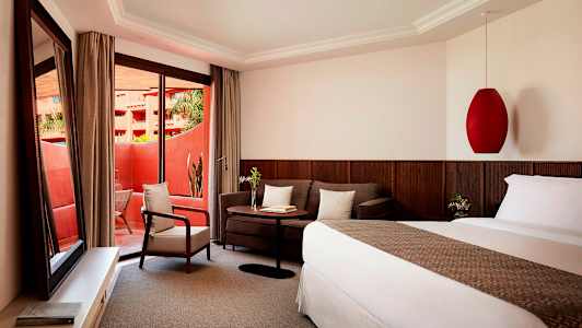 Ett hotellrum med en säng, en soffa, ett bord och en fåtölj. Rummet har stora glasdörrar som leder till en balkong. (AI-genererad text)