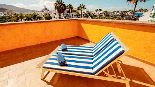 Utsikt från en solterrass med två solstolar med randiga blå och vita dynor samt handdukar på dem, omgivna av palmer och byggnader. (AI-genererad text)