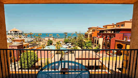 Utsikt från en balkong över en semesterresort med pooler, färgglada byggnader och havet i bakgrunden. (AI-genererad text)