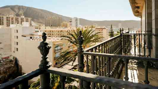 Balkong med detaljrik järnräcke och utsikt över stadsbyggnader med berg i bakgrunden. (AI-genererad text)