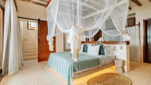 Ett sovrum med himmelssäng med vit myggnät, ljusa väggar och trägolv. En trappa syns i bakgrunden. (AI-genererad text)