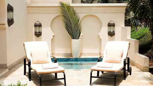 Två solstolar med dynor och kuddar står vid en pool framför en ljus vägg med dekorativa urtag och gröna växter. (AI-genererad text)