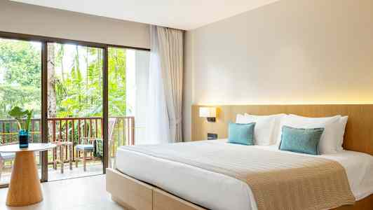 Ett hotellrum med en dubbelsäng, ljusa möbler och en balkong med utsikt mot trädgården. (AI-genererad text)