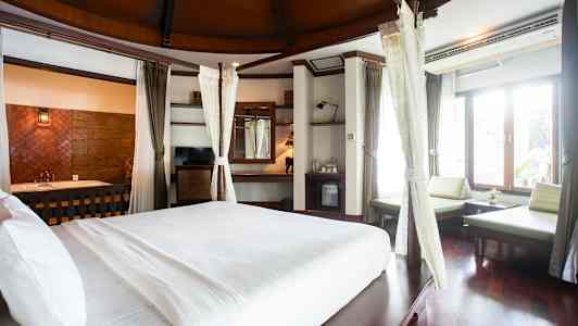 Ett rymligt hotellrum med en stor säng med vita lakan. Rummet har stora fönster som släpper in ljus, sittmöbler och trädetaljer. (AI-genererad text)